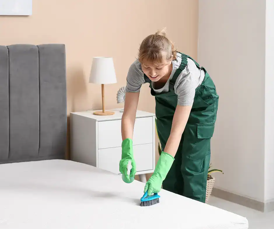 Cleaning specialist in green uniform scrubbing a mattress surface with a brush to remove stains in a modern bedroom