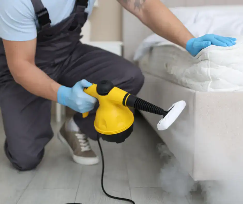 Professional technician in overalls using a handheld steam cleaner to deep clean and sanitize a white mattress in a bedroom
